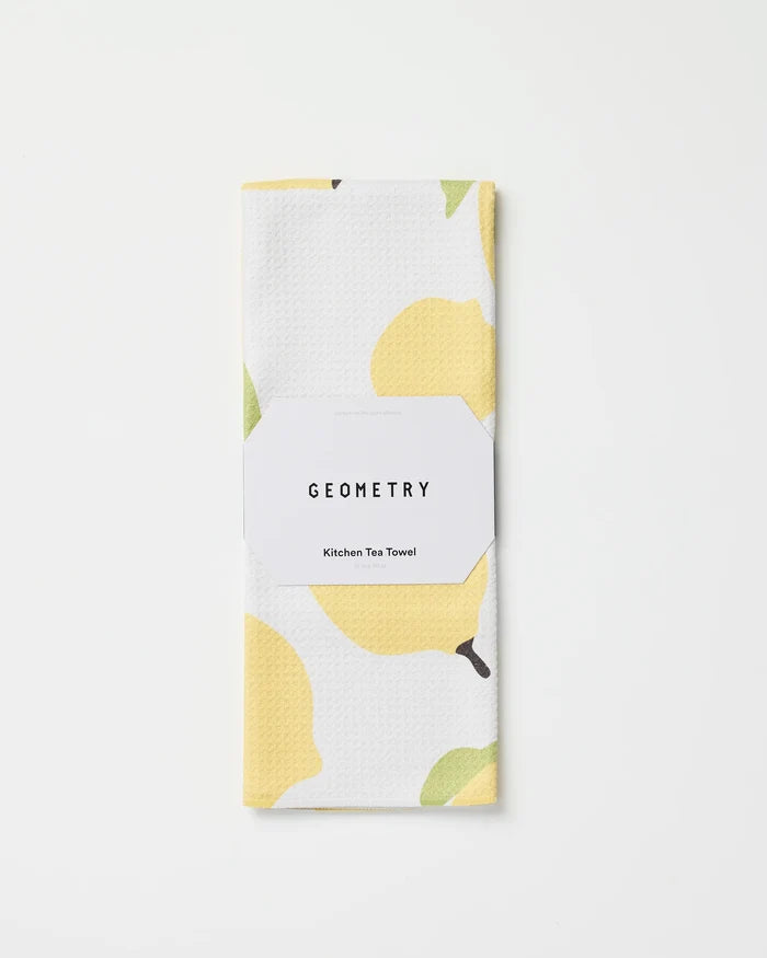 A folded white tea towel with a yellow lemon pattern, called Sunny Lemons by GEOMETRY, made from recycled materials and wrapped in branded packaging, rests on a plain white background.
