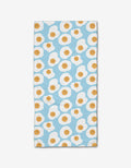 The Sunny Side Up bar towel by Janna Sue Design is rectangular, made from recycled materials, and features a light blue background with a repeating pattern of smiling cartoon fried eggs.