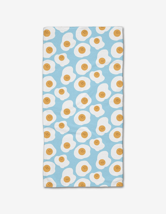 The Sunny Side Up bar towel by Janna Sue Design is rectangular, made from recycled materials, and features a light blue background with a repeating pattern of smiling cartoon fried eggs.