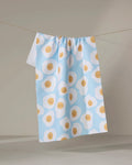 The "Sunny Side Up" tea towel by Janna Sue Design, crafted from recycled materials, features a blue background with sunny-side-up eggs. Ultra-absorbent and stylish, it brings sustainable charm to any kitchen.