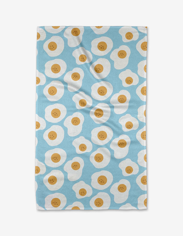 The Sunny Side Up tea towel by Janna Sue Design is blue, made from recycled materials, and displays a repeating sunny-side-up egg pattern with white and yellow details. Some wrinkles are visible when laid flat.