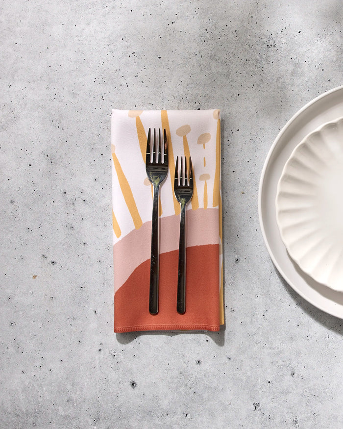 Two forks rest side by side on a folded GEOMETRY Sunny Stripes reusable dinner napkin with abstract patterns, placed on a light gray speckled surface next to a white scalloped plate.