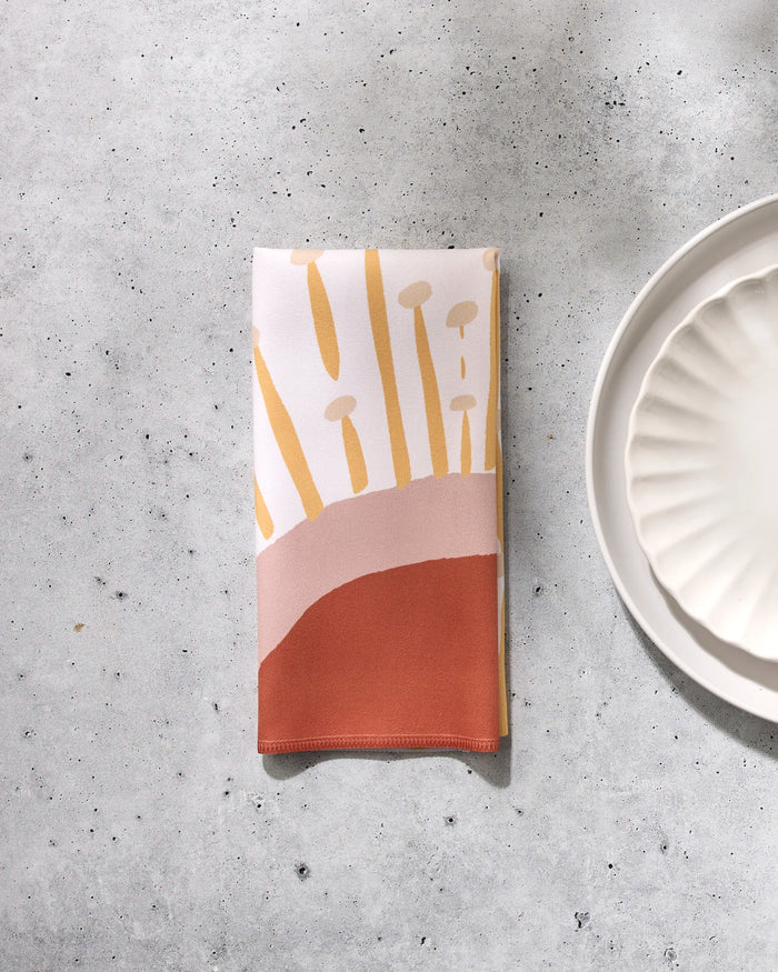 The GEOMETRY "Sunny Stripes" folded reusable dinner napkin, featuring abstract yellow, peach, and rust stripes, rests on a light gray speckled surface beside a white scalloped-edge plate.