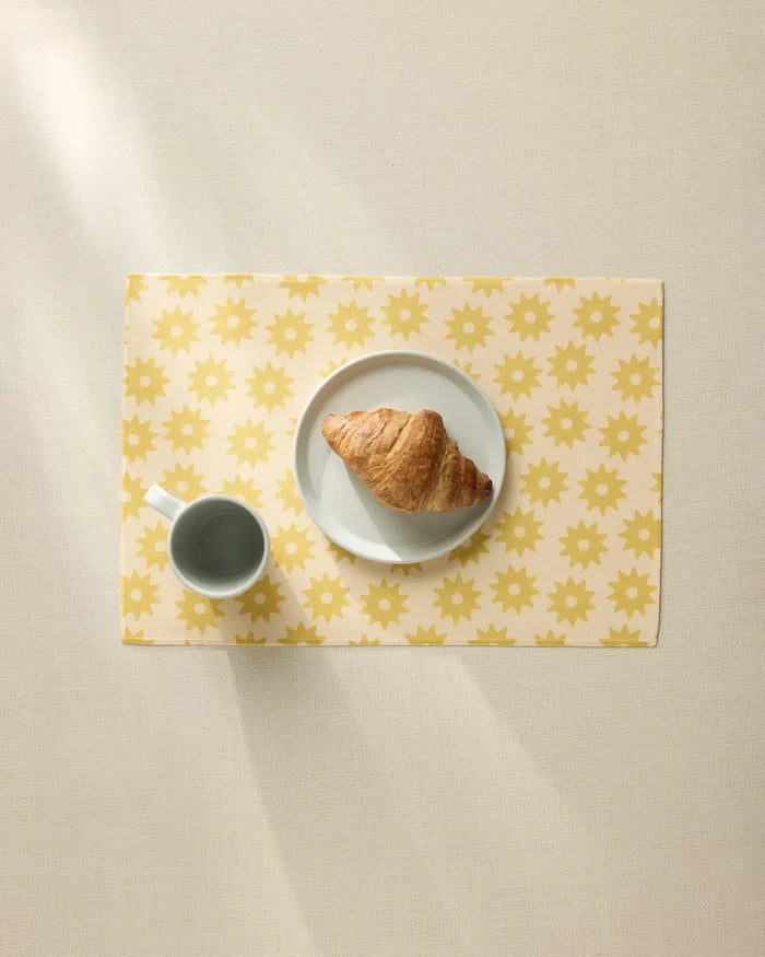A croissant on a white plate and a cup of coffee rest on the Geometry Sunny Sun placemat, featuring a yellow sun pattern, viewed from above on a light beige table.