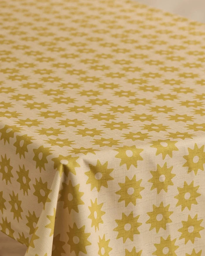 The Geometry Sunny Sun tablecloth, in beige with a repeating yellow sunburst pattern, covers a rectangular table.