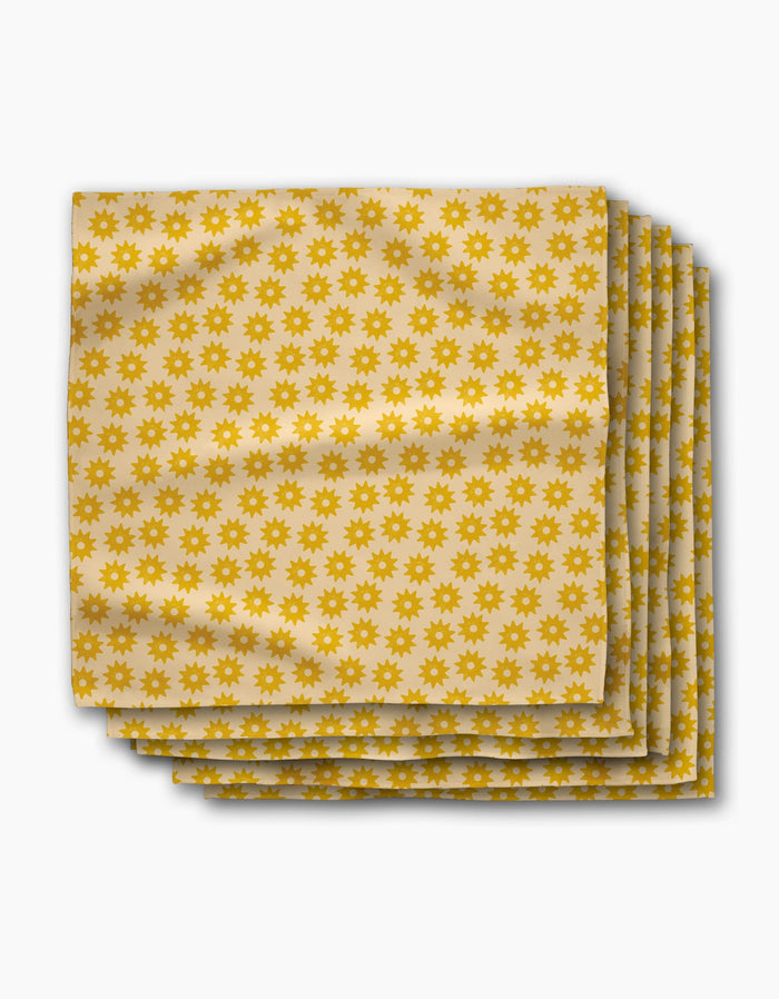 A stack of Sunny Sun beige eco-friendly napkins by Geometry, featuring a repeated yellow sunburst pattern, neatly arranged on a white background.