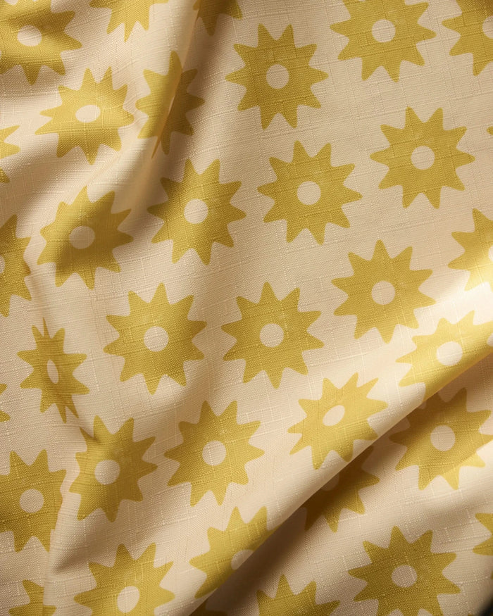 A close-up of Geometrys Sunny Sun tablecloth fabric in cream, featuring a lightweight design with yellow sunburst patterns, gentle folds, and a subtle textured weave.