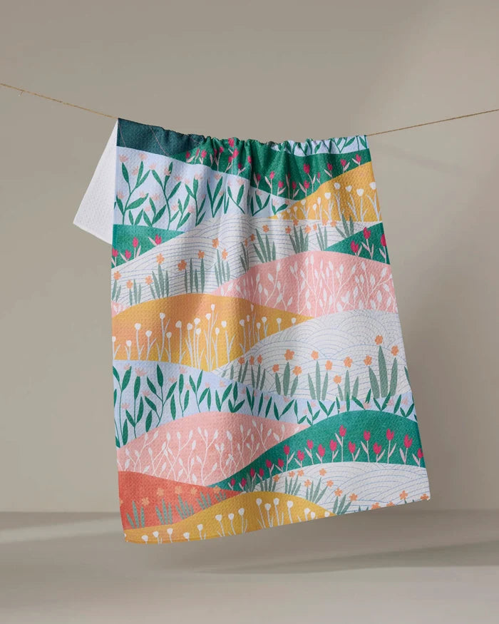 The Sunrise Mountains towel by Ceyda Alasar, featuring quick-drying fabric and vibrant wavy bands in green, yellow, pink, and orange with floral and grass motifs, hangs on a clothesline against a neutral backdrop.