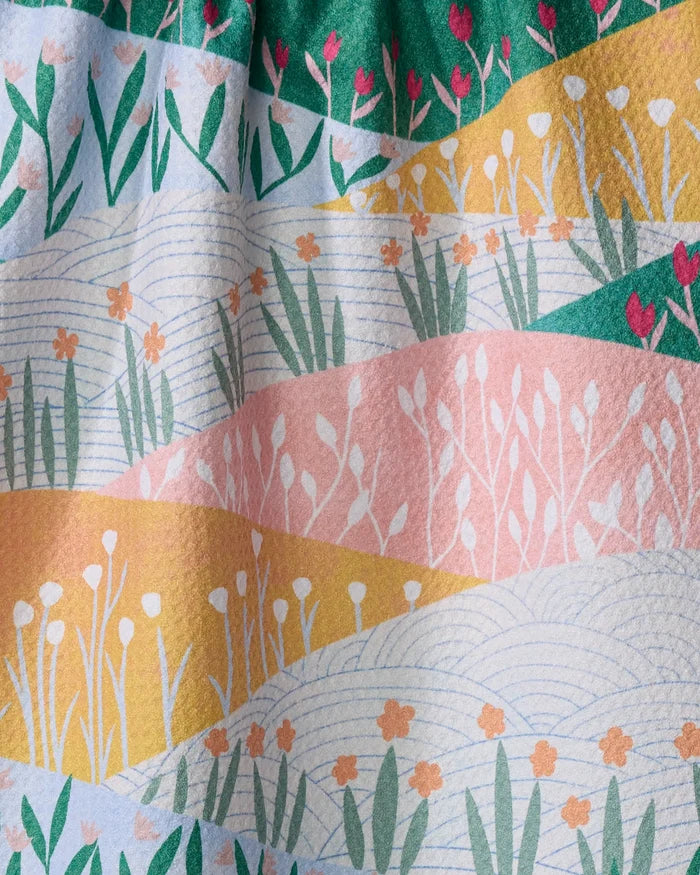 The "Sunrise Mountains" towel by Ceyda Alasar is a quick-drying towel with wavy stripes in green, yellow, pink, and white and stylized flowers and grass for a whimsical landscape look.