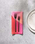 Two GEOMETRY Sunrise forks rest side by side on a folded eco-friendly napkin in pink and orange, next to a white scalloped plate atop a light gray speckled surface.