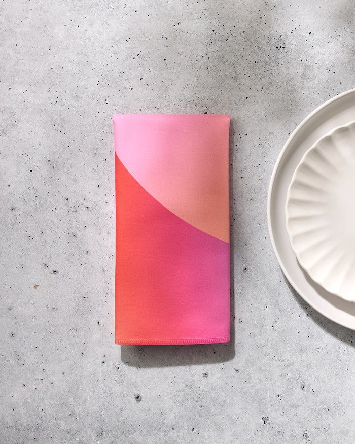 The Sunrise eco-friendly napkin by GEOMETRY, featuring a pink and orange gradient, is folded on a light speckled surface beside a scalloped white plate, with soft shadows completing the scene.