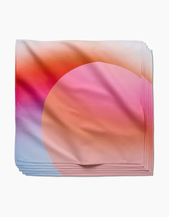 Sunrise by GEOMETRY is a folded square scarf with a gradient in pink, orange, purple, and light blue hues, crafted from recycled materials and featuring a large semi-transparent peach circle on smooth fabric.