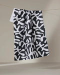 Sunset" by Marina Solodka, a recycled kitchen towel featuring a bold black and white abstract pattern of organic shapes, hangs on a line against a plain beige background.
