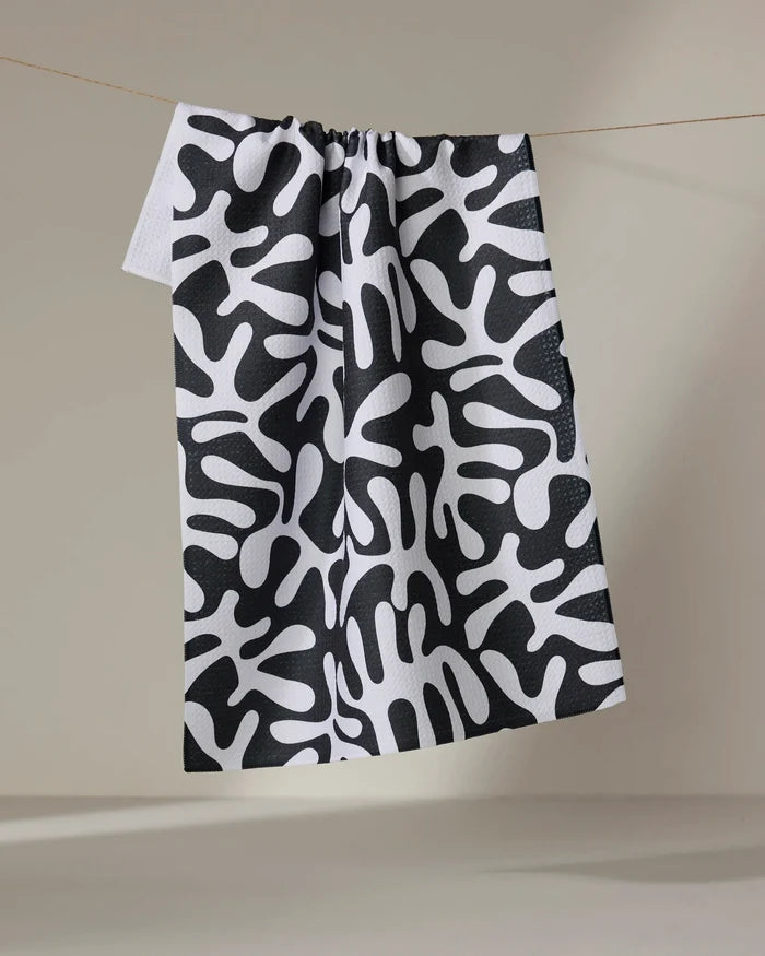 Sunset" by Marina Solodka, a recycled kitchen towel featuring a bold black and white abstract pattern of organic shapes, hangs on a line against a plain beige background.