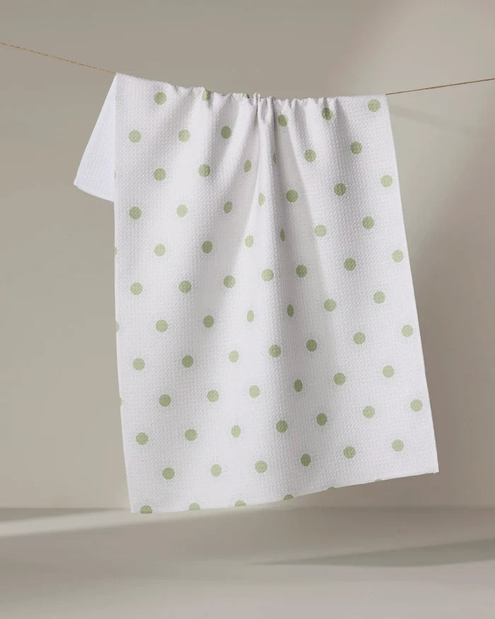 The Danelys Sidron "Sunshine Glow" tea towel, white with a textured surface and light green polka dots, hangs on a clothesline against a neutral background.