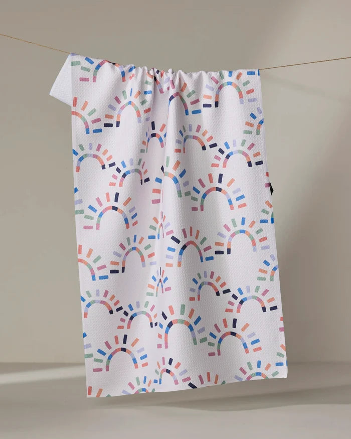 The "Sunshine Rainbow" tea towel by Claire Iglesias features colorful, semi-circular rainbow patterns and hangs on a line against a neutral background.