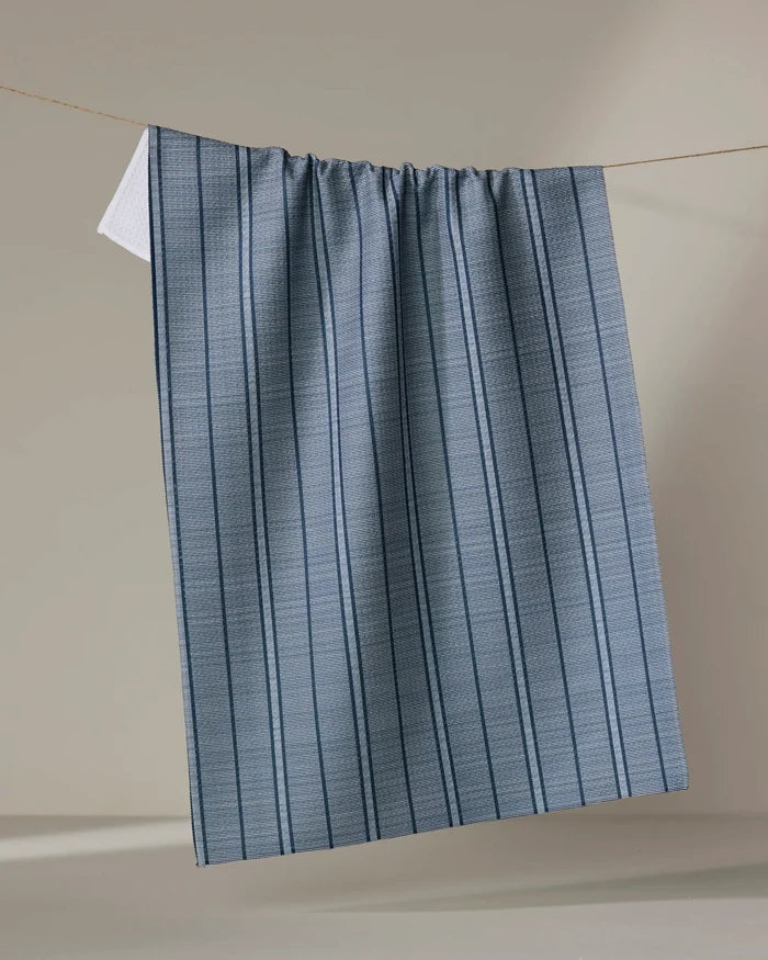 The Buy Guide’s Super Stripe - Navy kitchen towel, made from recycled materials, hangs from a thin line with blue and gray vertical stripes and a subtle texture—perfect as an absorbent dish towel against a neutral beige background.