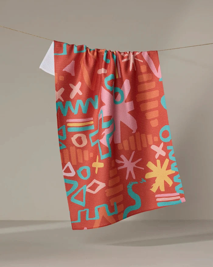 The Sareka Unique "Superstar" towel features playful geometric and squiggle patterns in turquoise, yellow, and pink on a vibrant red-orange base, quick-drying and eye-catching, shown hanging against a plain background.