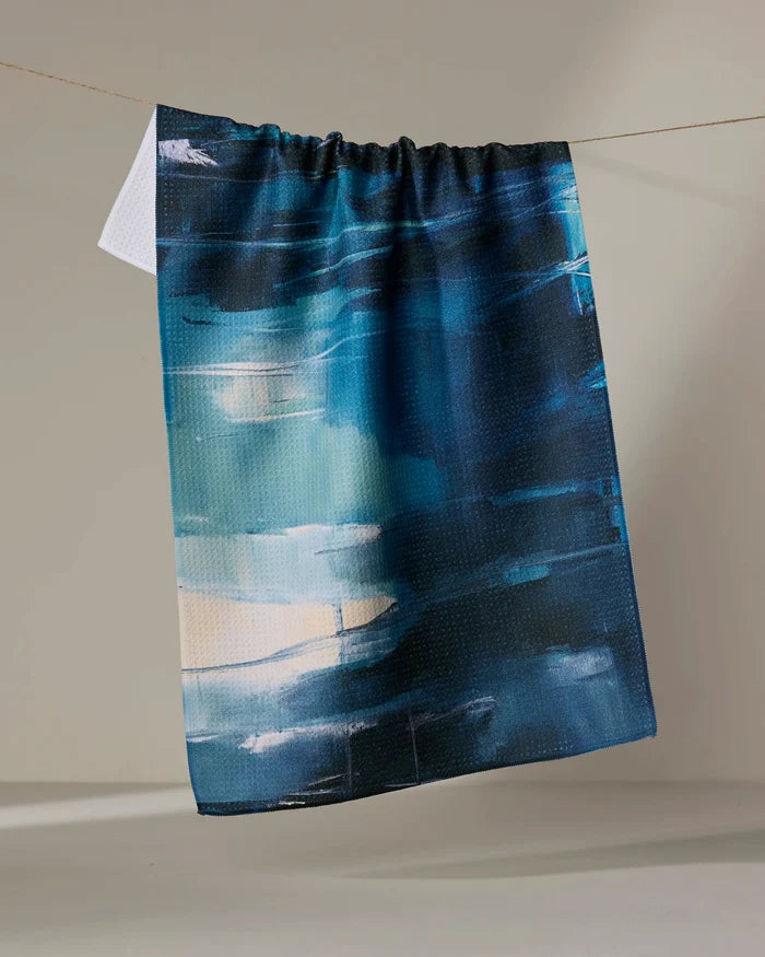 The "Surface Tension" tea towel by Jennifer Janesko, featuring an abstract design in blue, teal, and white, hangs on a thin line against a plain light beige background.