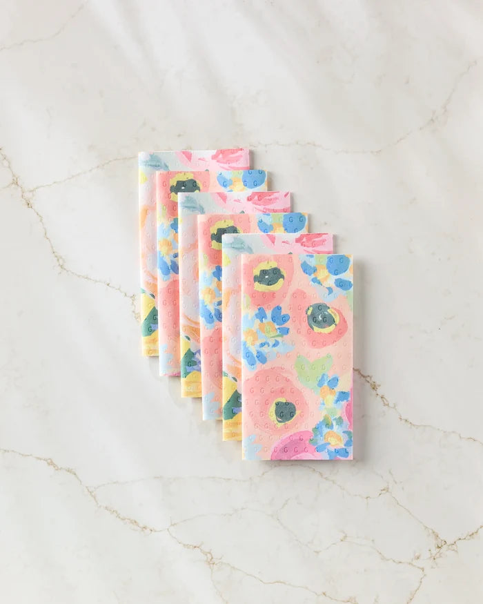 Six eco-friendly Suzzy and Margot paper towels by Ania Zwara Art are neatly stacked on a white marble surface. The napkins display pastel pink, blue, and yellow flowers with green leaves.