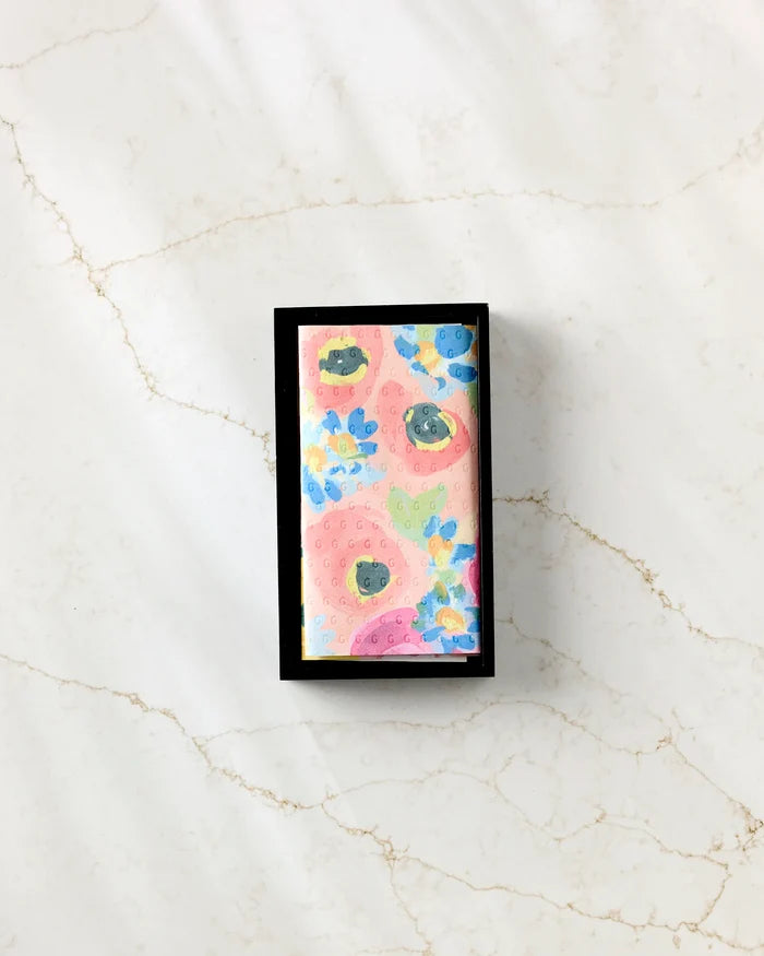 The Suzzy and Margot bar soap by Ania Zwara Art features a colorful abstract floral design in blue, green, and pink. It comes in a black box, displayed on white marble—an elegant touch next to eco-friendly paper towels.