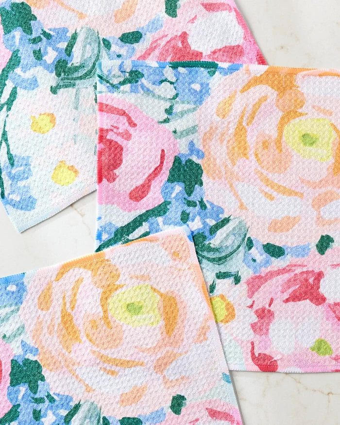 The Suzzy dishcloth set by Ania Zwara Art features four recycled cloths with textured surfaces and vibrant abstract floral patterns in pink, yellow, blue, green, and orange, arranged overlapping on a light marble background.