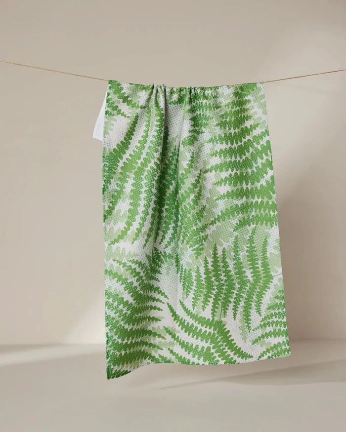 The Swaying Leaf tea towel by GEOMETRY, featuring a green and white repeating fern motif and made from recycled materials, is displayed hanging on a thin string against a neutral beige background.