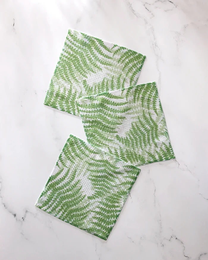 Three square green and white GEOMETRY Swaying Leaf dishcloths, featuring a delicate wavy leaf pattern, are arranged in a staggered line on a white marble surface.