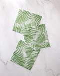 Three square green and white GEOMETRY Swaying Leaf dishcloths, featuring a delicate wavy leaf pattern, are arranged in a staggered line on a white marble surface.