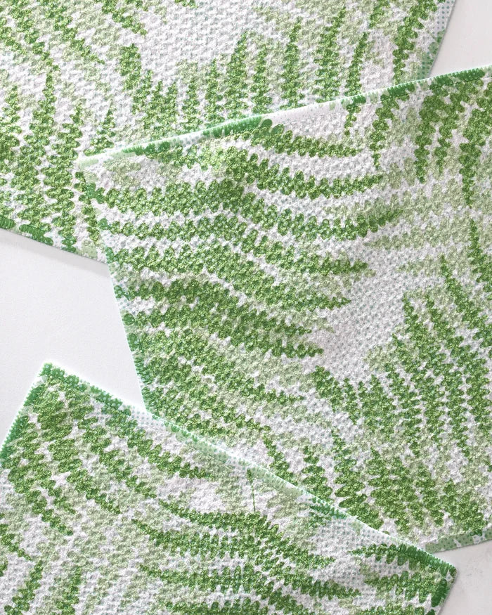 Four GEOMETRY "Swaying Leaf" dishtowels, featuring white fabric with green wavy leaf-like patterns, overlap on a white surface, creating a textured, abstract look.