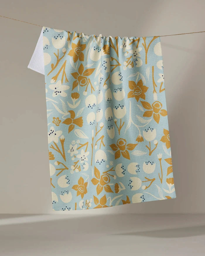 The "Swedish Cherry Blossoms" kitchen towel by Elisabeth Wing features a gold and white floral and berry pattern on light blue, made from recycled materials, hanging on a clothesline against a neutral background.