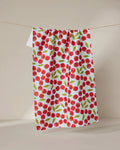 The GEOMETRY Sweet Cherry quick-drying tea towel, featuring a red cherry and green leaf pattern, hangs on a thin string against a plain beige background.