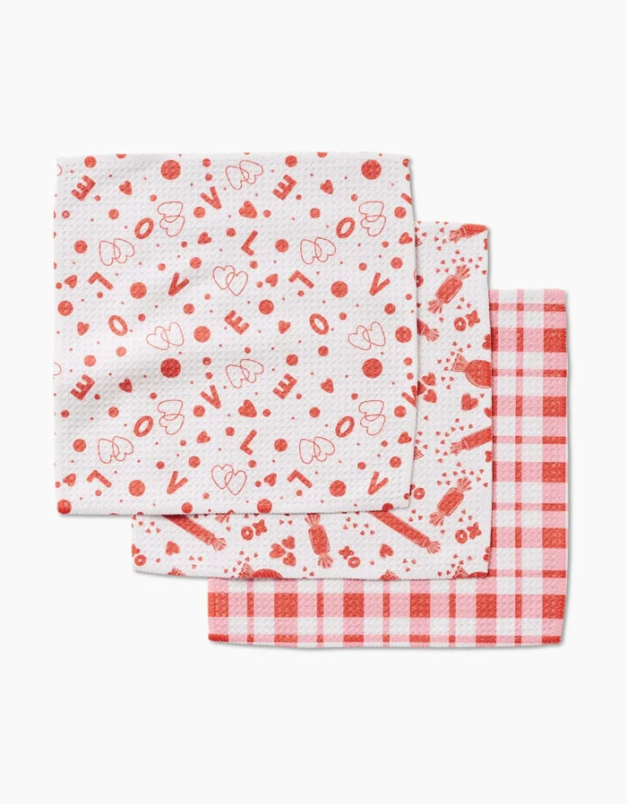 Three folded Sweet Love dishcloths by Charlie Rowan Designs—one with hearts and “Love” text, one with strawberries and dots, and one red-white checkered—are neatly stacked, all made from recycled materials.