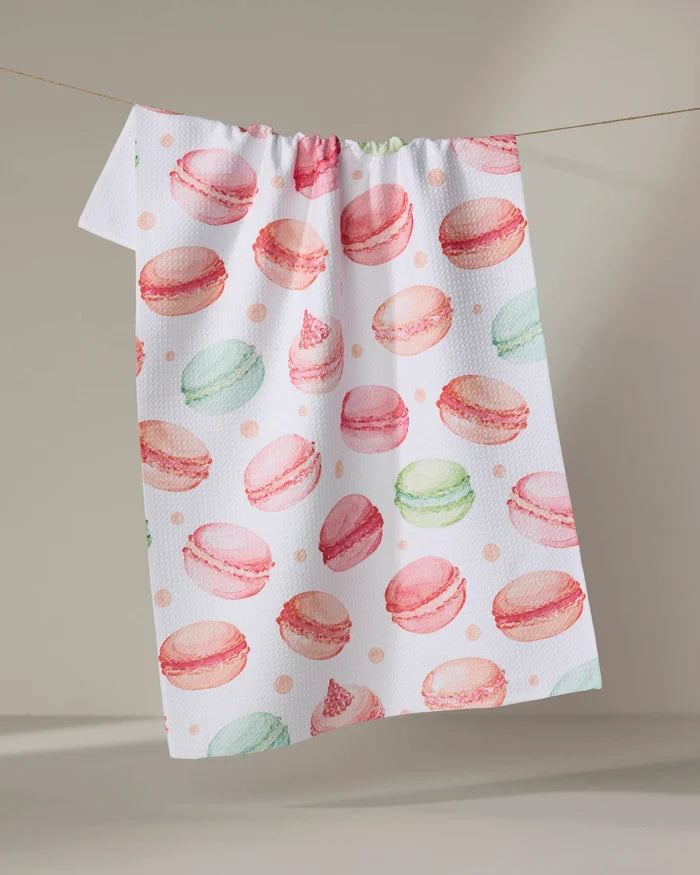 The Elena Fay "Sweet Macarons" white tea towel, crafted from recycled materials and adorned with a vibrant pink, green, and peach macaron print, hangs on a thin string against a neutral background.