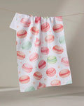 The Elena Fay "Sweet Macarons" white tea towel, crafted from recycled materials and adorned with a vibrant pink, green, and peach macaron print, hangs on a thin string against a neutral background.