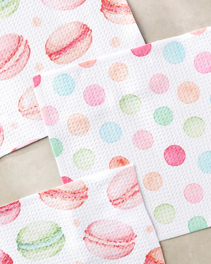 Three textured napkins from Elena Fay's "Sweet Macarons" set lie on a light surface: two with colorful macaron illustrations, and one with pastel polka dots—an eco-friendly choice made from recycled materials.