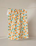 The GEOMETRY Sweet Orange tea towel features a vibrant orange fruit and green leaf pattern on white, hanging against a neutral backdrop—a stylish, quick-drying addition to any kitchen.