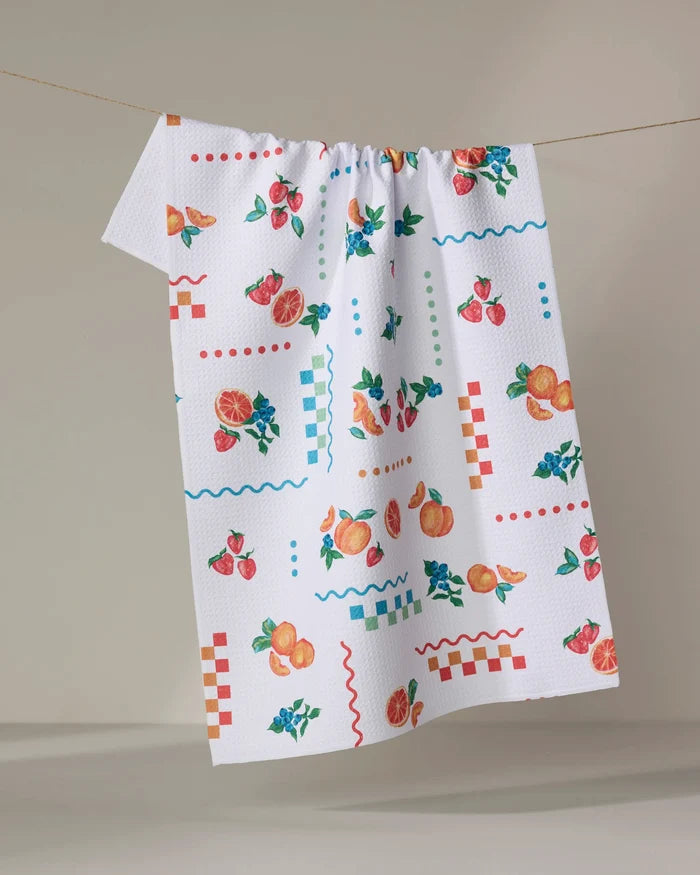 The Sweet Pickings tea towel by Aubrey Fairchild features colorful fruit illustrations—like oranges, strawberries, and peaches—and red and blue checkerboard patterns, hanging on a clothesline against a neutral backdrop.