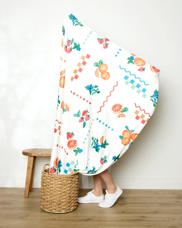 A person stands behind the Sweet Pickings Butter Blanket by Aubrey Fairchild, featuring fruit and geometric patterns—only their legs and white sneakers visible—beside a woven basket and wooden bench against a white wall.