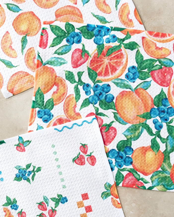 The Sweet Pickings set by Aubrey Fairchild features three super-absorbent dishtowels with vibrant fruit patterns—orange, strawberry, blueberry, and peach—adding a colorful touch to your kitchen decor.