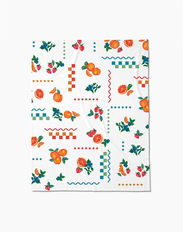 The Aubrey Fairchild Sweet Pickings Butter Blanket offers silky softness with a vibrant design of oranges, strawberries, leaves, and colorful geometric shapes in red, blue, and green.