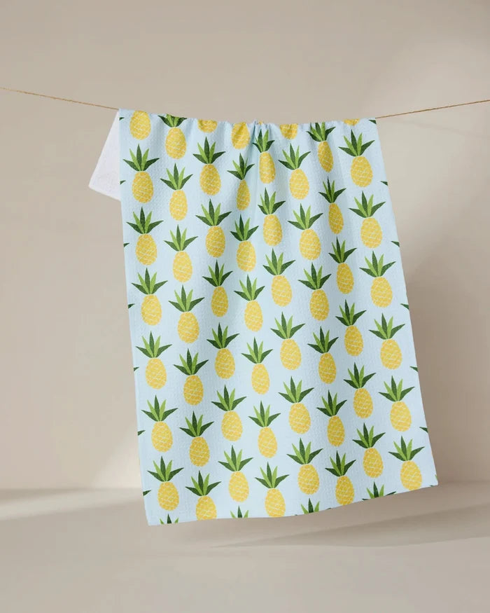 The Sweet Pineapple tea towel by GEOMETRY, featuring a light blue color with a repeating pattern of yellow pineapples and green leaves, hangs from a thin line against a neutral beige backdrop.