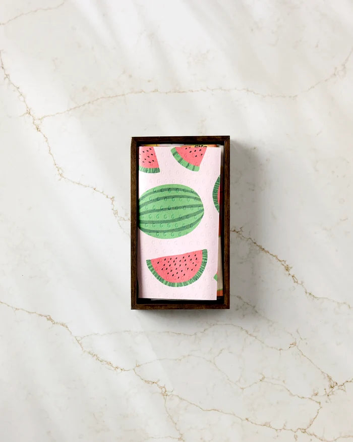 The GEOMETRY Sweet Summer Mix 2 pink napkin, decorated with watermelon illustrations and resembling eco-friendly paper towels, rests neatly inside a rectangular wooden holder on a light marble surface.