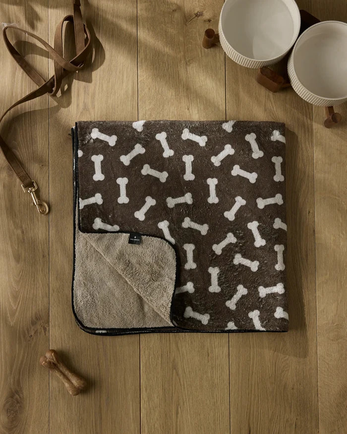 The Geometry "Sweet Treats" plush dog towel in brown with a white bone pattern lies on a wooden floor, next to two white bowls, a brown leash, a dog bone toy, and two wooden cubes.