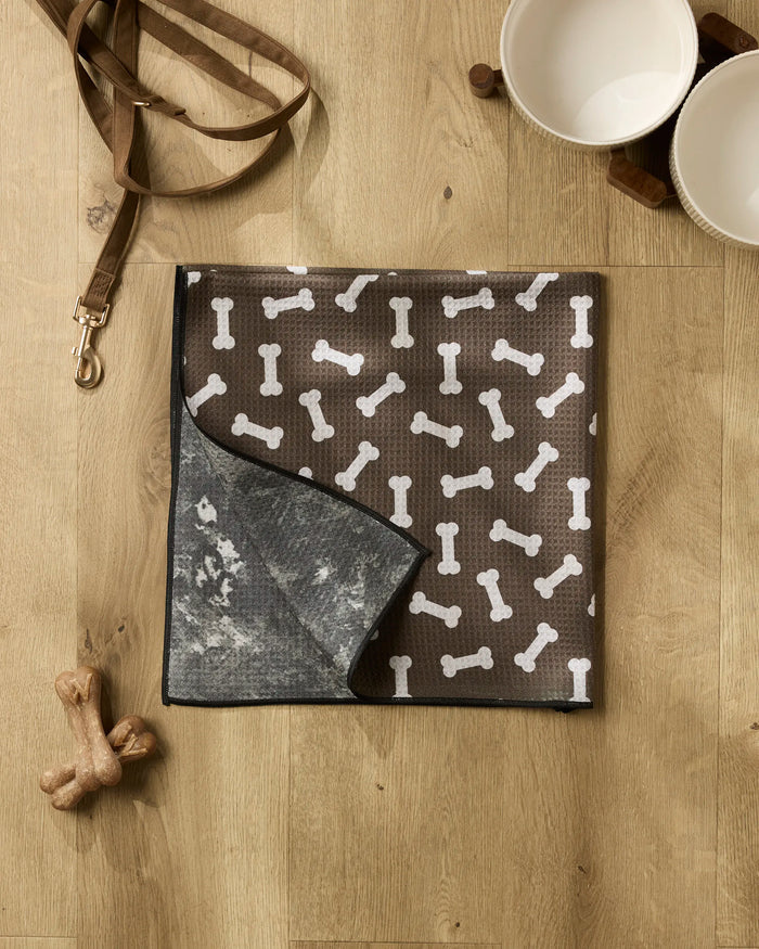 A brown dog mat with white bone patterns from Geometry's Sweet Treats collection lies on a wooden floor, joined by a recycled dog towel, leash, two ceramic bowls on a wooden stand, and two dog bone toys for a cozy corner.