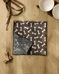 A brown dog mat with white bone patterns from Geometry's Sweet Treats collection lies on a wooden floor, joined by a recycled dog towel, leash, two ceramic bowls on a wooden stand, and two dog bone toys for a cozy corner.