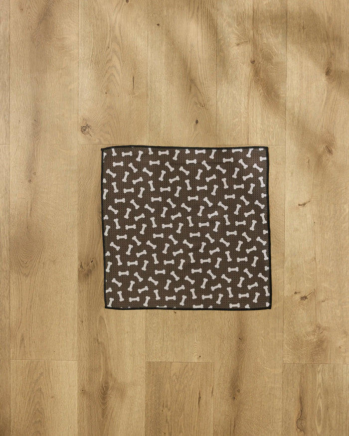 The Geometry Sweet Treats mat, featuring a brown square design with white dog bone patterns, sits on a light wood floor and doubles as an ultra-absorbent dog towel to keep your pet’s paws dry and clean.