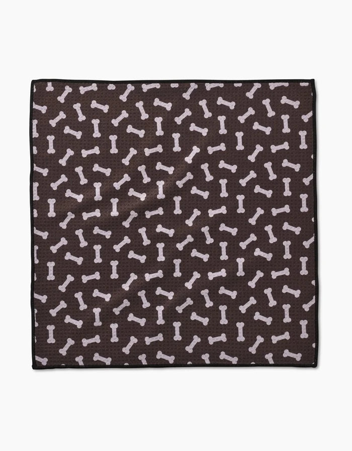 The Sweet Treats Paw Towel by Geometry is a square, dark brown towel made from recycled materials, featuring a repeating pattern of light-colored dog bones in various directions.
