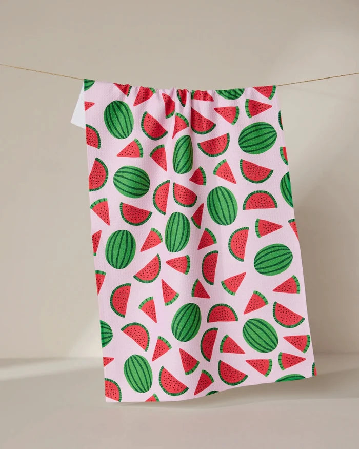 The GEOMETRY Sweet Watermelon tea towel, featuring colorful watermelon slice and whole fruit designs on white, hangs on a string against a neutral background.