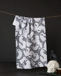 The Geometry Swirling Ghosts tea towel, featuring a cartoon ghost pattern, hangs against a dark background with a white skull and stacked books nearby for a spooky Halloween vibe.